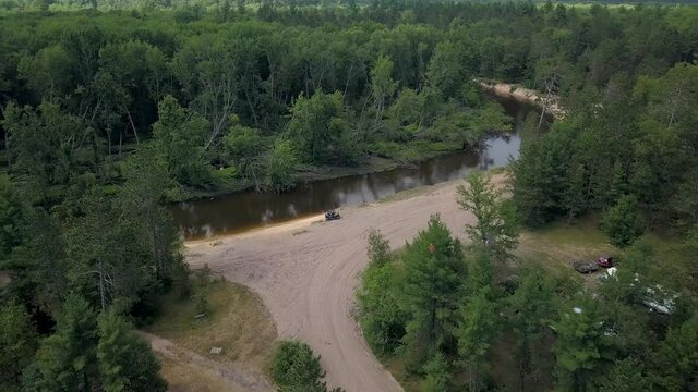 4k Drone Flyby Unrecognizable Man In Side By Side UTV On A River Side ORV Nature Trail In Mid Michigan With Sunny Skies Muddy Water And Campers And Trails All Around