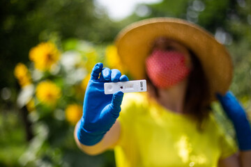 Woman in yellow top wearing a red facemask and hat holding a coronavirus rapid test in front of sunflowers bokeh