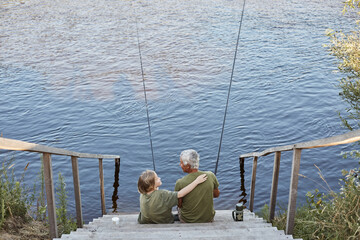 Fototapeta premium Happy family spending time together in open air near river or lake, son hugging his father with love while sitting on wooden stairs leading to water.
