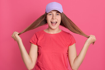 Fototapeta na wymiar Positive young lovely female pulling her long straight hair with raised hand and looking cheerfully directly at camera, isolated over pink background.
