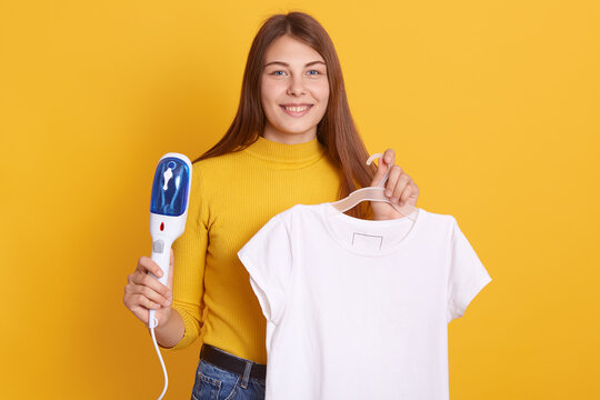 Young woman being ready to steam her clothes on rack at home, preparing for dating, wearing yellow casual shirt, looking smiling directly at camera.