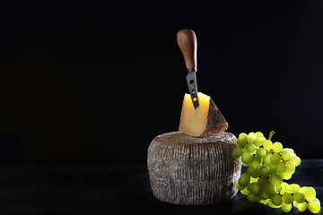 A whole head of hard craft cheese. A piece of cheese with a knife. A branch of green grapes nearby. Photo on a black background. Copy of the space.