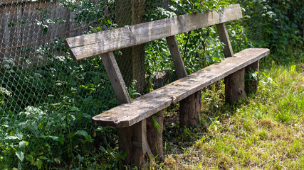 Wooden bench in the village, against the background of green bushes and a fence made of metal mesh netting. Intimate relaxation in the countryside in the fresh air.