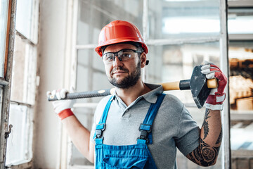 Worker, looking out the window, stands at the construction site. He is wearing a red safety helmet and he holds a sledgehammer for repairs in his hands.