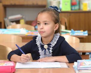 A junior schoolgirl sits at a desk and thinks. The student looks to the side holding the pen in her right hand. A girl in a school uniform thought about sitting at a table.