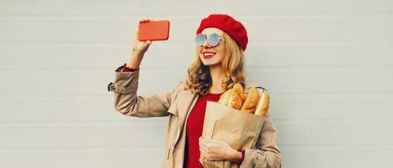 Portrait of smiling woman taking selfie picture by phone holding grocery shopping paper bag with long white bread wearing red beret over gray background