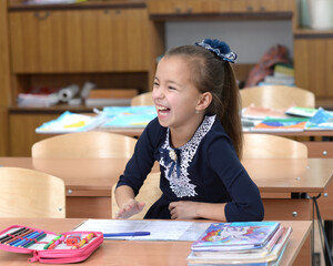 A primary school student laughs at a desk. The schoolgirl sits at the table and laughs. A girl in a blue school uniform is a lot of fun.