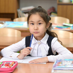 A junior schoolgirl sits at a desk and thinks. The student holds a pen near the chin. The blue pen in the hands of the schoolgirl. A girl in a school uniform thought about sitting at a table