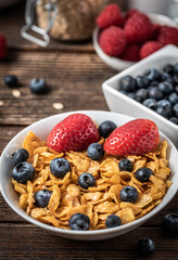 Corn flakes with blueberries and raspberries in white bowl in dark wooden desk.
