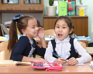 Elementary school students sit at the desk and secret. A schoolgirl whispers secrets in the ear of her school friend. The girl is surprised by listening to his friend.