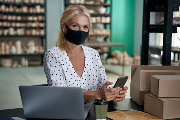 Female business owner wearing protective face mask holding mobile phone and looking at camera while working in her art studio or craft pottery shop with handmade ceramic products