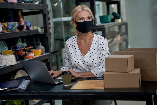 Small business after lockdown quarantine. Female business owner or business woman wearing protective face mask working on laptop in her art studio or craft pottery shop
