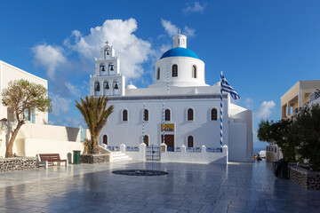 Santorini, Oia - View of the white bell tower, in the background a church with a blue dome in the foreground of a cobbled, stone square.