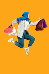 Jumping high. Young muslim woman with shopping packages on yellow studio background. Stylish, trendy. Beautiful brunette. Human emotions, facial expression, sales, ad, shopping concept. Copyspace.