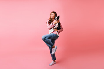 Full-length portrait of adorable girl in trendy jeans holding french bulldog. Dreamy caucasian woman posing with her dog on pink background.
