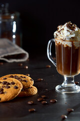 Close up of a glass cup of coffee with whipped cream and chocolate on it, chocolate chip cookies and roasted coffee beans on dark background. Concept of ready to eat food, tasty snack. Selective focus