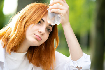 Young tired thirsty redhead woman drinking water from a bottle in summer park.