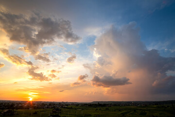Dramatic sunset over rural area with stormy clouds lit by orange setting sun and blue sky.