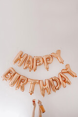 Female hands hold rose champagne bottle and glass. Rose gold balloons Happy Birthday on white wall. Holiday anniversary party celebrating decoration