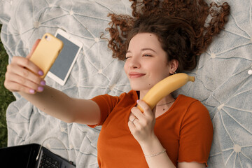 Close up lifestyle portrait of young smiling girl lying on the ground and making selfie while holding banana in hand. Woman taking picture with the help of modern gadget and blinking.