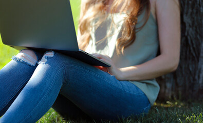 Girl sitting on the green lawn in the park, typing and working on laptop computer. Freelance outside 
