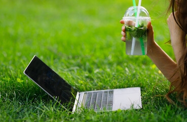 Image of girls hand with a lemonade works with a laptop in the park on a green lawn.