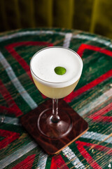 A classic yellow sour cocktail with foam in a nick and nora glass with lime zest garnish