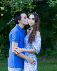 Vertical portrait of cute young man in blue shirt kissing his pretty girlfriend’s cheek with eyes closed while standing in garden during a summer late afternoon, Quebec City, Quebec, Canada 