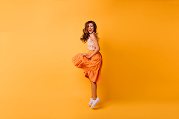Fototapeta na wymiar Full-length portrait of romantic beautiful lady in orange skirt. Stylish carefree girl jumping on yellow background.