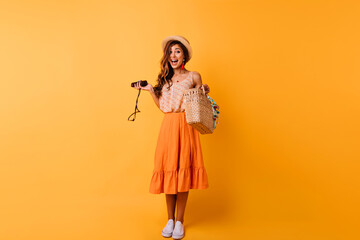 Full-length portrait of sensual woman wears white shoes and straw hat. Well-dressed caucasian girl making funny faces on orange background.