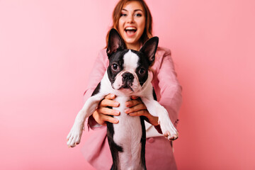 Portrait of stylish carefree girl with little funny dog on foreground. Charming caucasian lady with dark hair expressing good emotions during photoshoot with french bulldog.