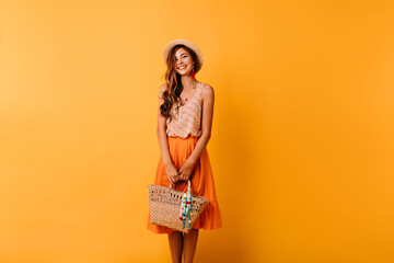 Elegant woman in summer outfit preparing for vacation. Romantic ginger girl in straw hat posing on orange background with bag.