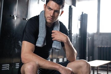 Young athletic Caucasian man sitting alone in dark gym locker room resting after workout