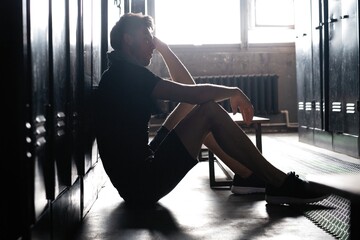 Fototapeta na wymiar Young athletic Caucasian man sitting alone in dark gym locker room resting after workout