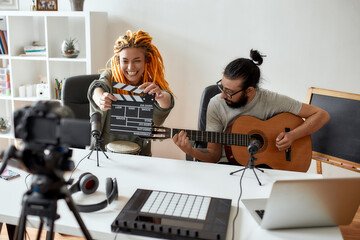 Face the music. Man playing guitar and woman holding clapperboard. Couple of musicians recording video blog or vlog while making music at home