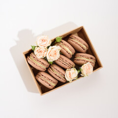 Many chocolate macarons in craft box with roses on the white background.