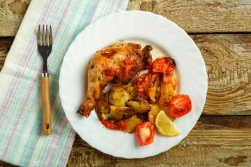Baked chicken leg with potatoes and tomato on a white plate with a fork on a wooden table.