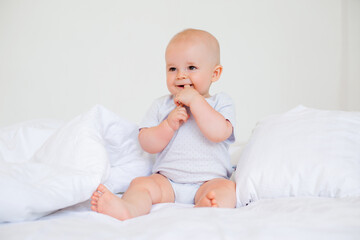 Cute baby boy 6 months smiling in a white bodysuit sitting on a bed on white bedding