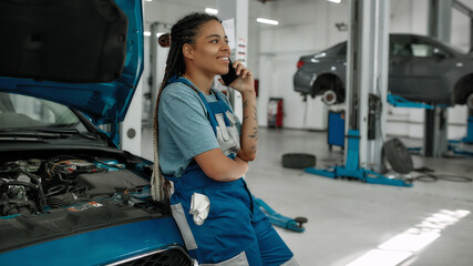 Young african american woman, professional female mechanic talking on phone, leaning on a car with...