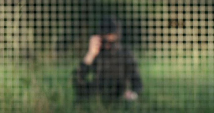 A man in a black mask threatens by phone while sitting on the grass behind bars, blurred background.