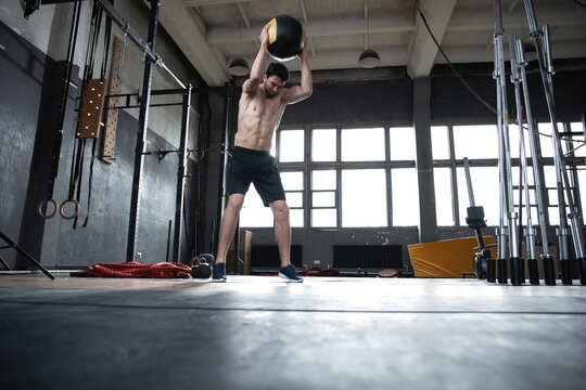 Fit man throwing medicine ball doing ball slam against gym floor or shoulder press upper body workout exercise. Cross training at fitness center.