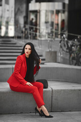 Obraz premium Young woman with wearing stylish red suit sits on the bench outdoors on the city street