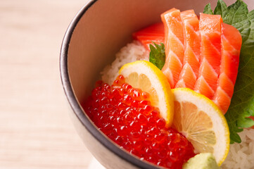 raw pink salmon sashimi and red salmon roe ikura over white rice with lemon wedges in a ceramic bowl