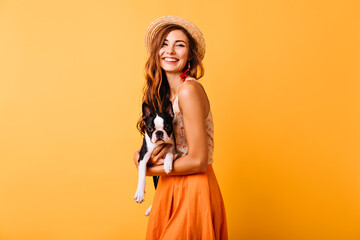 Stunning ginger girl in summer outfit holding french bulldog. Appealing young lady in hat laughing during photoshoot with puppy.