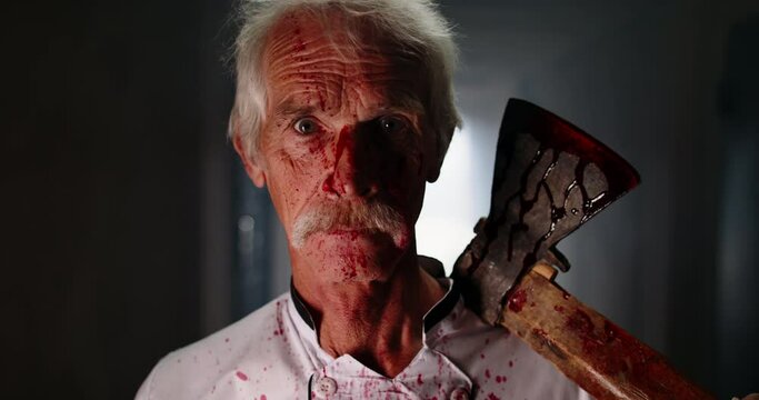 Mustached senior caucasian psycopath creepily staring at camera and grinning, wearing butcher's apron covered in blood and carrying bloodstained axe - halloween concept 4k portrait