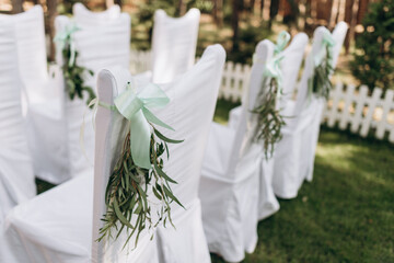 rows of white chairs for the wedding ceremony are decorated with green willow branches and ribbons. wedding outdoors ceremony