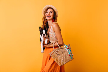 Glad woman in summer hat holding french bulldog. Laughing girl in orange skirt posing with funny puppy.