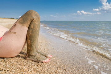 View of beautiful female feet with bright red pedicure on the sand of the beach. The ankles of the feet are smeared with healing mud. Skin rejuvenation while relaxing at sea. Spa. Copy space.