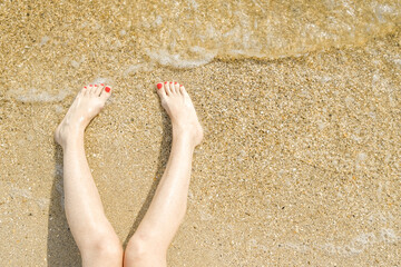 Top view of beautiful female feet with bright red pedicure on the sand of the beach. The sea wave washes women's feet. Relaxation and enjoyment during your seaside holiday. Copy space