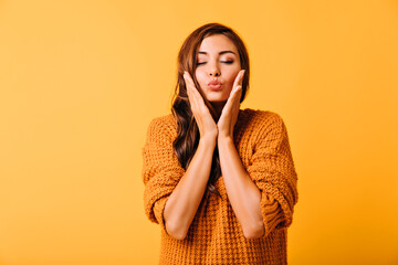 Dreamy girl in knitted clothes touching her face. Inspired brunette woman with light makeup standing on yellow background.
